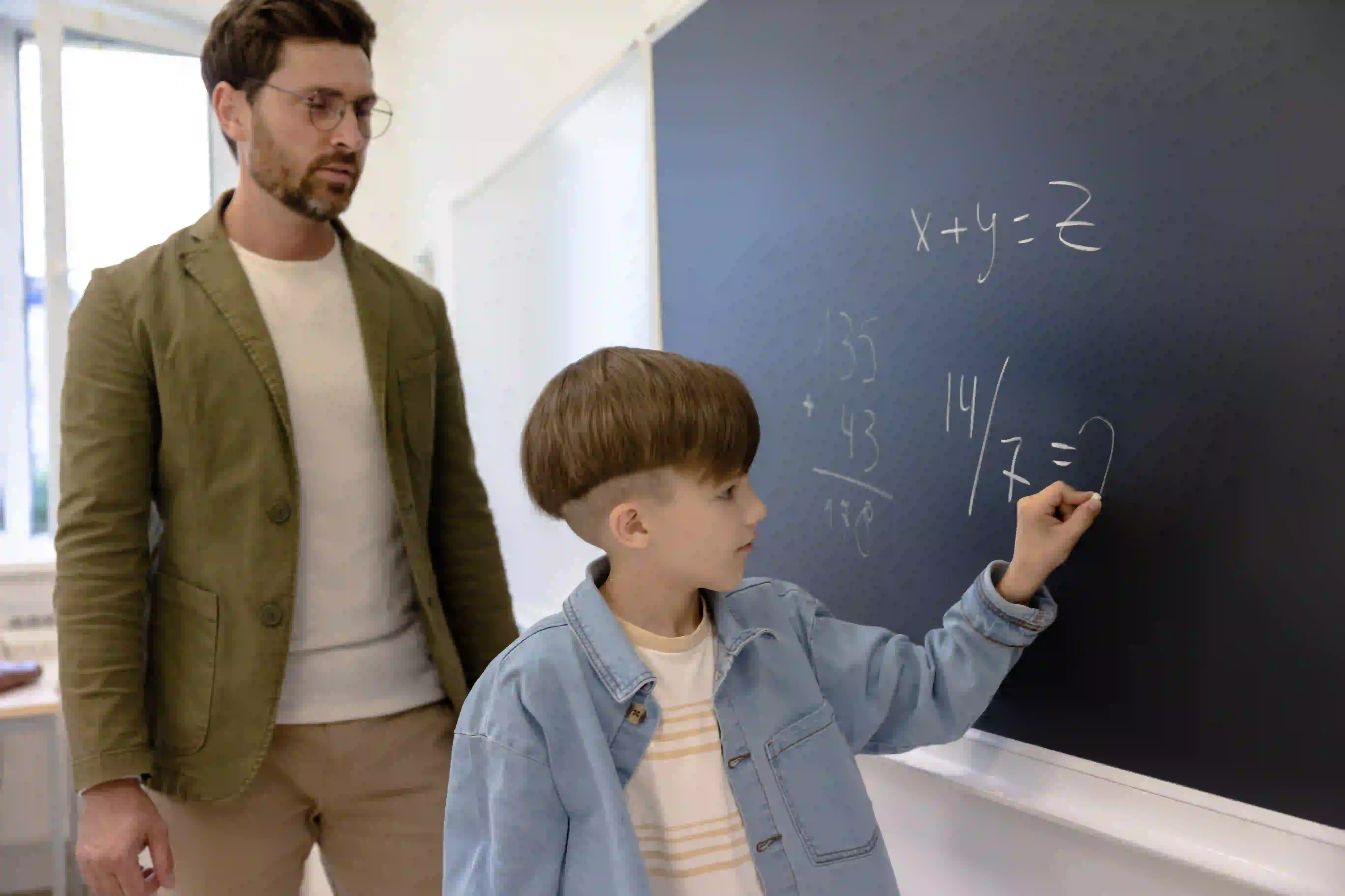 Grundschüler löst eine Matheaufgabe an der Tafel, während der Lehrer daneben steht und zuschaut.