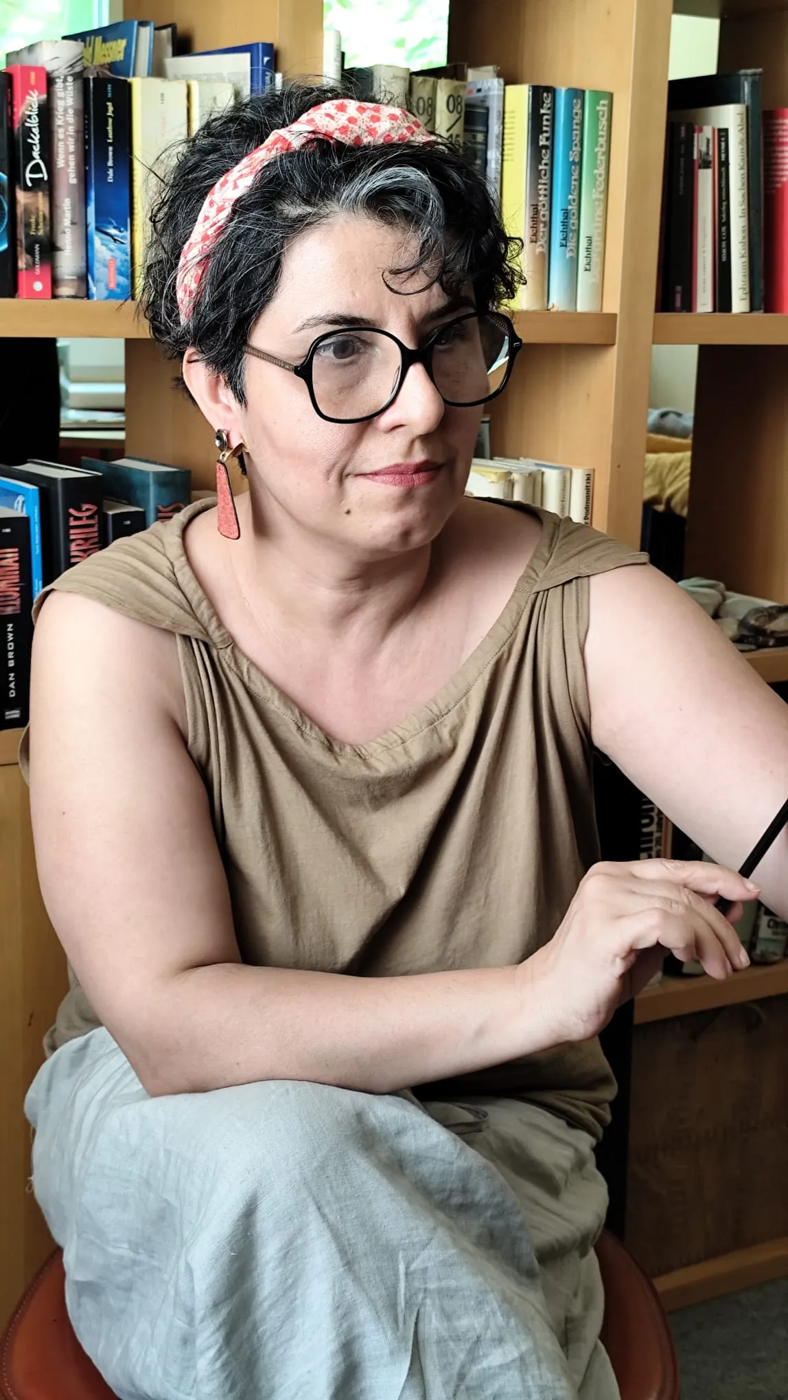 Frau mit kurzen dunklen Haaren und Brille sitzt vor einem Bücherregal, trägt ein beigefarbenes Oberteil, eine Leinenhose und ein rotes Haarband mit Muster.